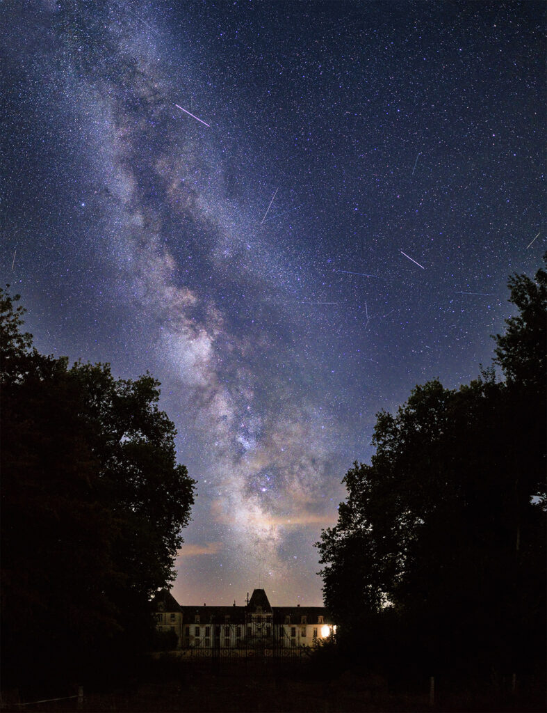 Starry night sky with the Milky Way and several meteors, framed by dark trees and a lit building at the bottom.