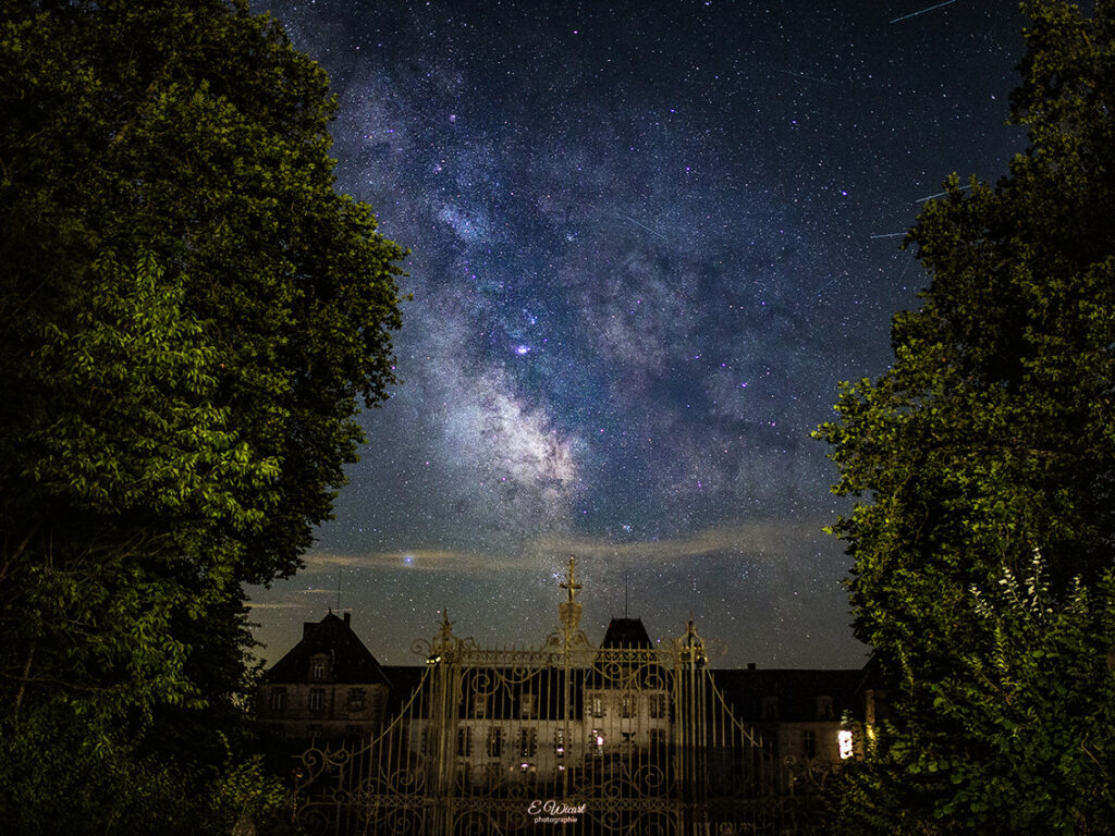 Milky Way arcing across a starry night above an ornate gate and a castle, framed by trees in the foreground.