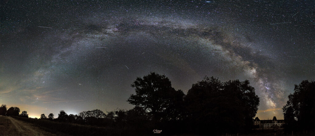 Wide night sky with the Milky Way arc over a rural landscape; silhouettes of trees along a dirt road beneath a star-filled sky.